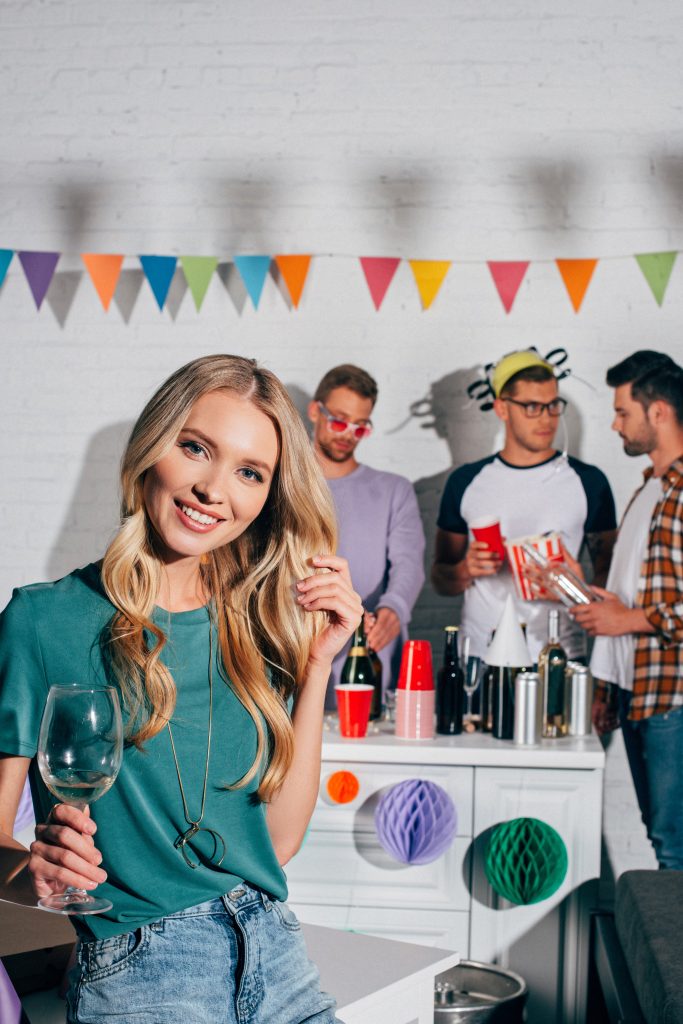 Jonge vrouw in turquoise shirt met wijnglas poseert lachend op een feest met vrienden en gekleurde slingers op de achtergrond.