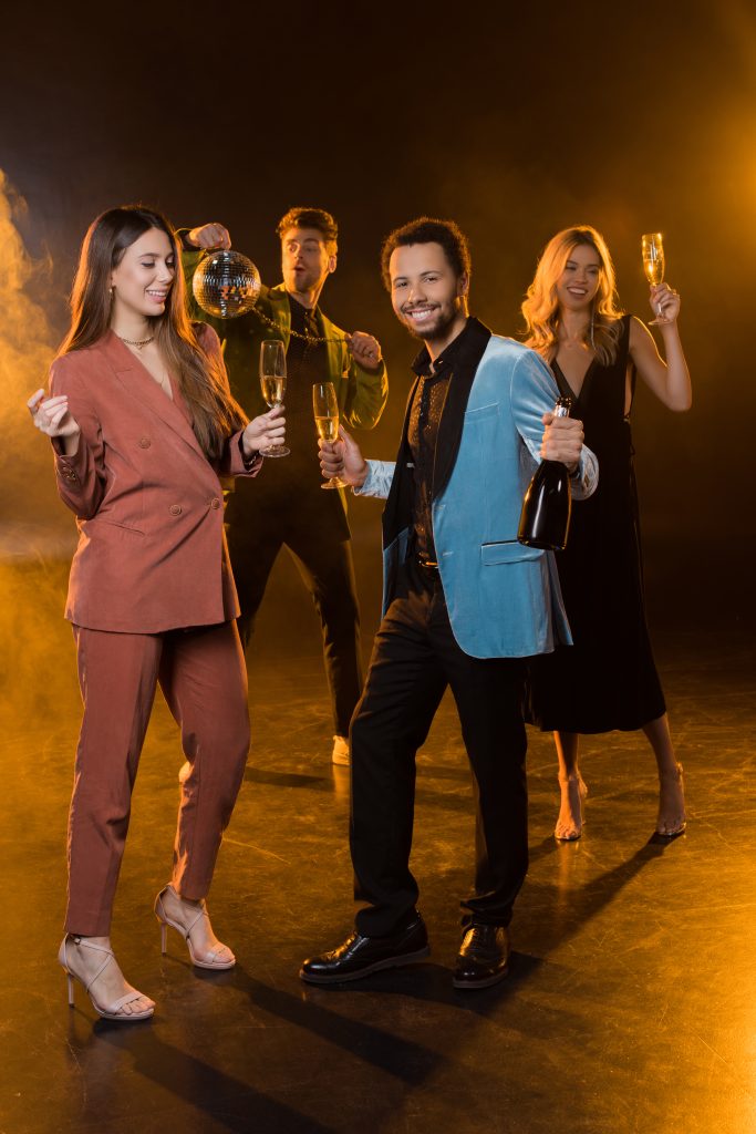 Vijf feestende mensen in elegante kleding houden champagneglazen en een discobal omhoog tegen een gouden achtergrond.