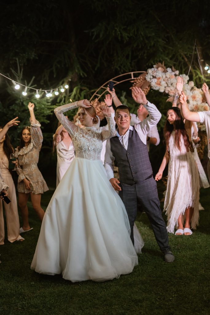 Bruidspaar danst onder bloemen en versiering in avondtuin, omringd door feestende gasten met opgeheven armen.