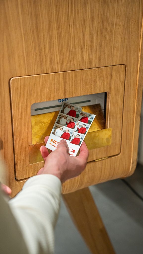 Hand haalt fotobewijs uit houten DNP-fotoautomaat met gele uitvoerbak.