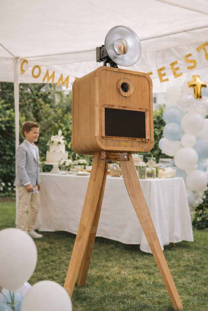 Houten fotocabine op driepoot met flitser op een communie-feest in een tuin onder witte tent.