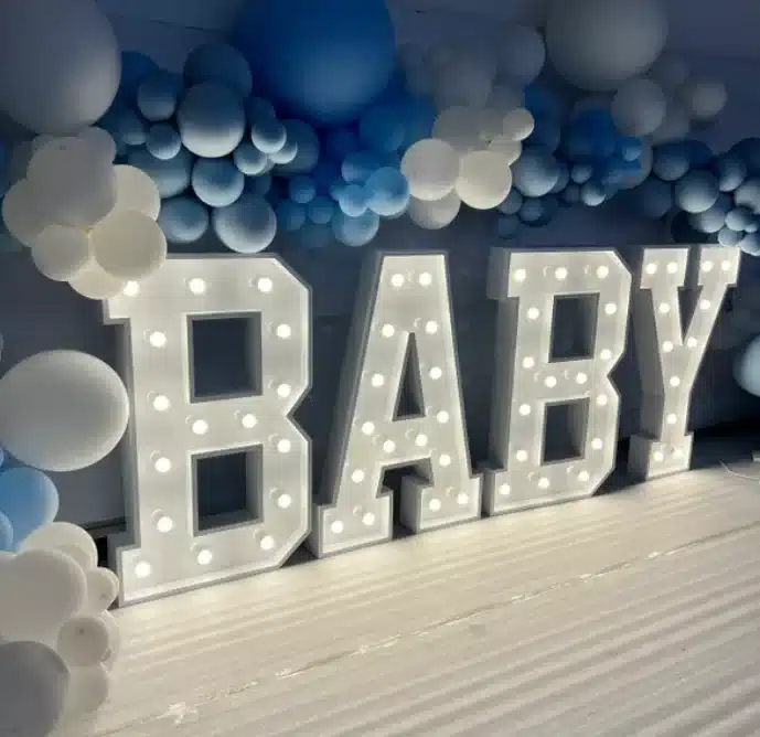 Verlichte houten letters spellen "BABY" en staan op een houten ondergrond met blauwe en witte ballonnen erboven.