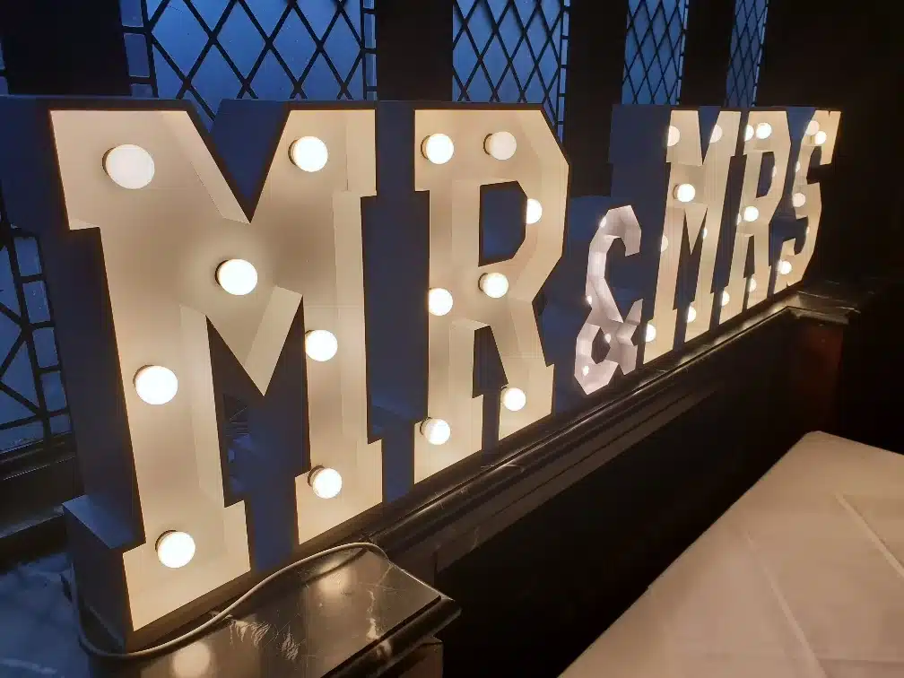 Verlichte lichtletters met witte gloeilampen spellen "MR&MRS" op een zwarte basis tegen een blauwe achtergrond.