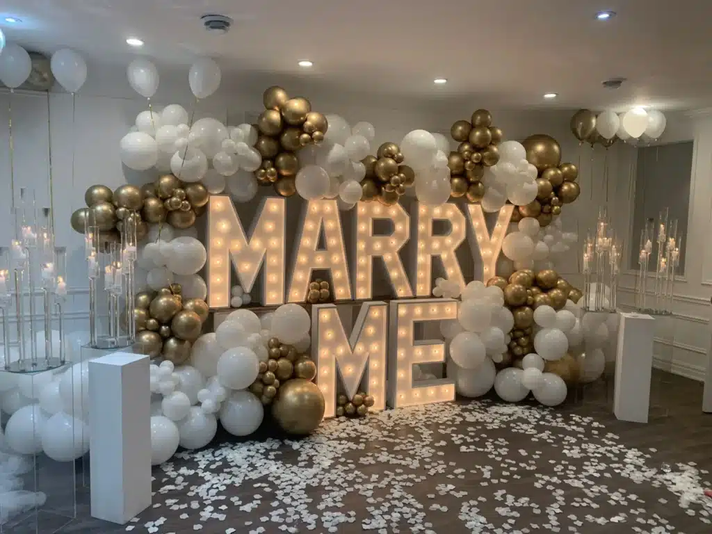 Verlichte letters spellen "MARRY ME" omgeven door witte en gouden ballonnen in een elegant ingerichte ruimte voor huwelijksaanzoeken.