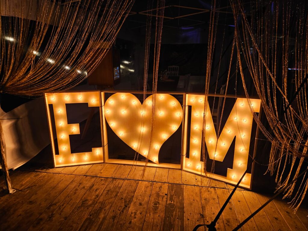 Drie verlichte lichtletters, een E, een hart en een M, op een houten vloer tijdens een trouwfeest.
